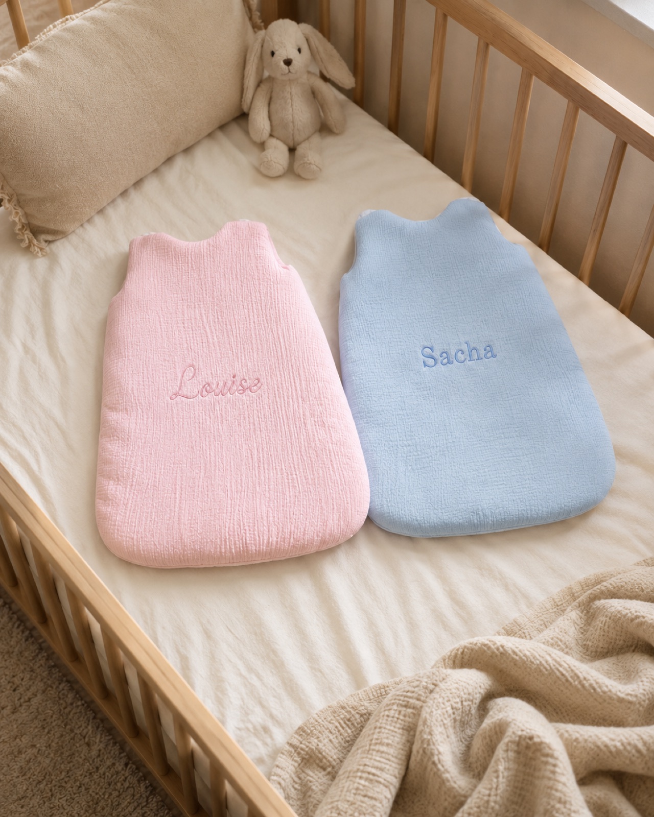 Deux gigoteuses personnalisées NuBaby, rose poudré et bleu ciel, brodées aux prénoms, posées dans un lit à barreaux