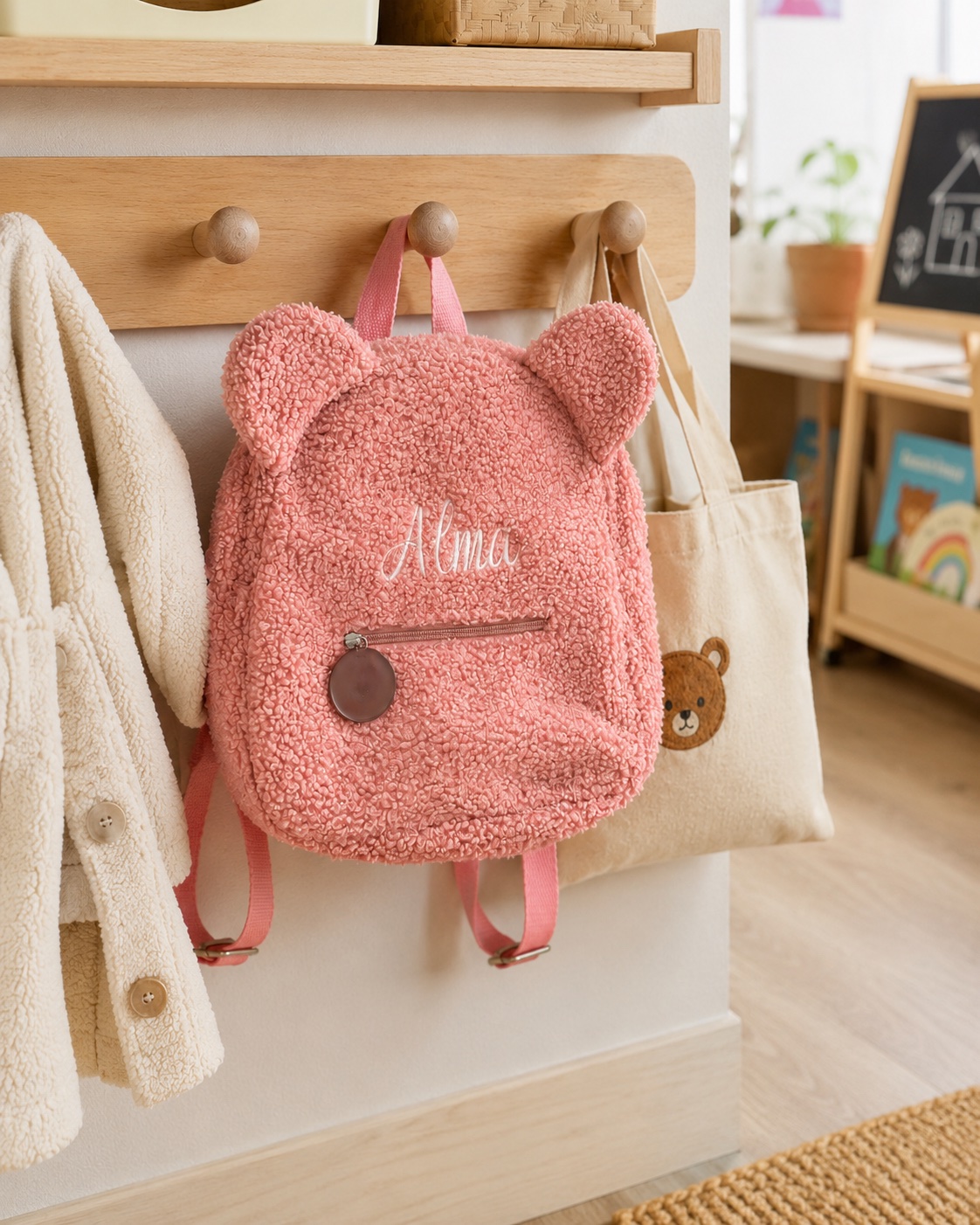 Petit sac à dos rose effet bouclette à oreilles d’ourson, prénom brodé, suspendu à une patère dans une entrée